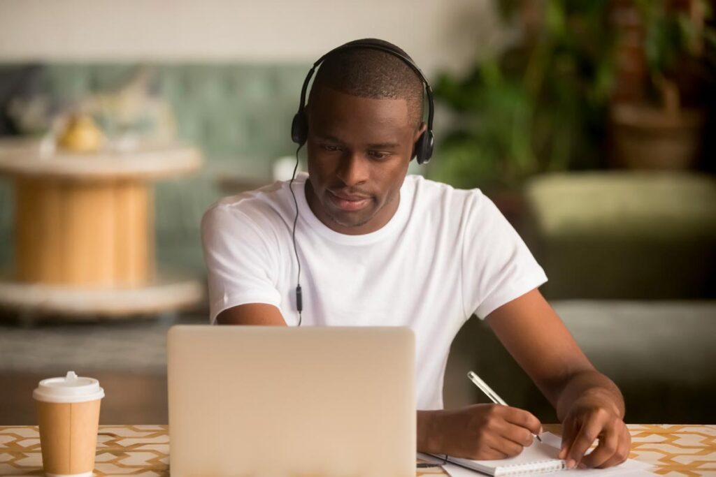Young adult man wearing headphones, writing on notepad while doing research on laptop